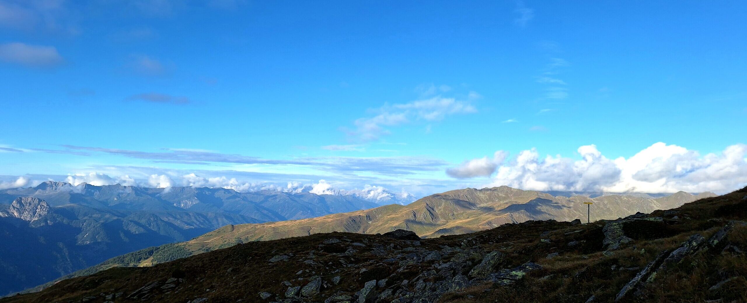 Gebirgspanorama unter strahlend blauem Himmel, leichte Bewölkung im Hintergrund, fern rechts in der Bildmitte ein Wegweiser